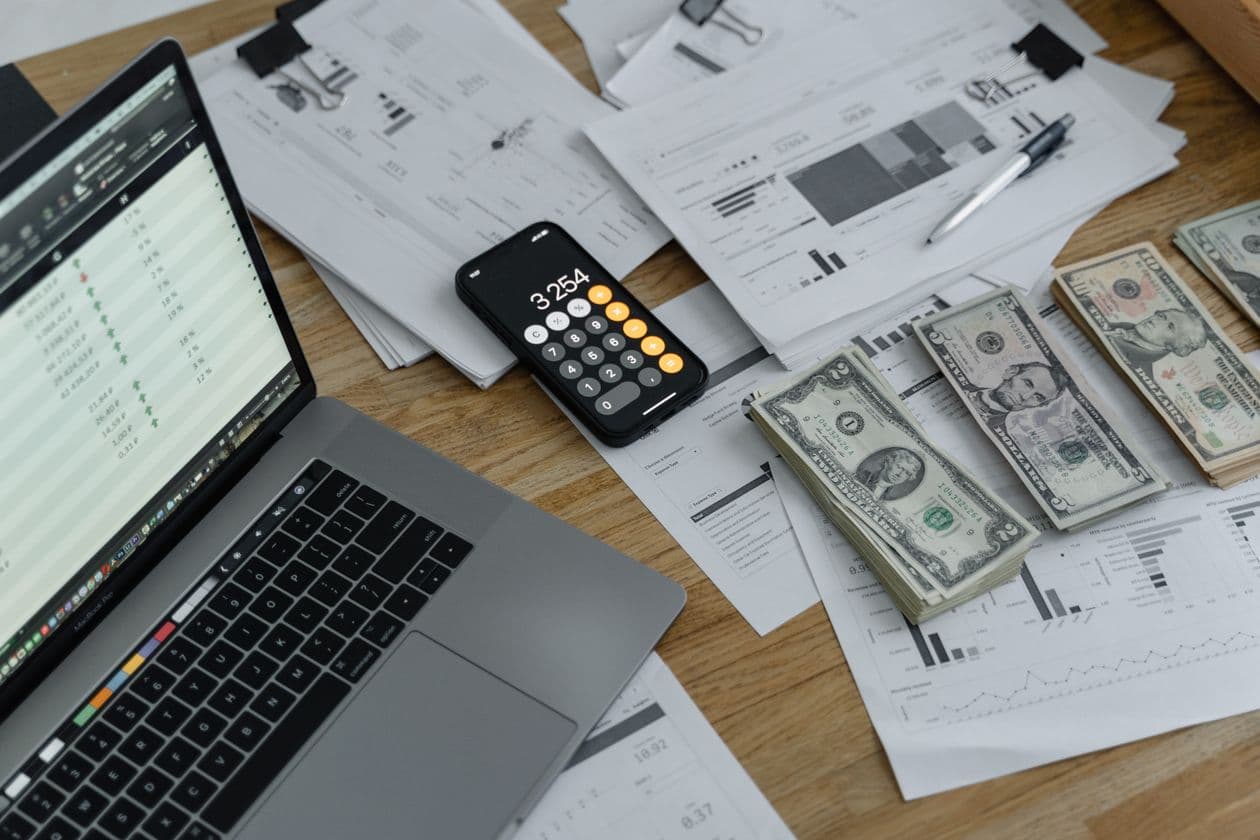 Laptop with a spreadsheet next to a calculator, cash, and printed reports